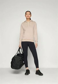 Gai bēšs sporta krekls ar kapuci un auklu, melnas legingi un melnas sporta kurpes. Tur pie rokas melna sporta soma. Vienkāršs dizains, minimāla zīmola klātbūtne.