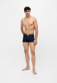 Мъж стои бос на бял фон, облечен в тъмносини боксерки Polo Ralph Lauren.