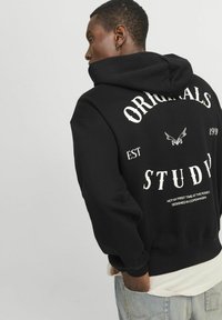 Zwarte hoodie met witte tekst op de achterkant, met grote "ORIGINALS" en "STUDY", en een klein logo. Zachte stof, buidelzak.