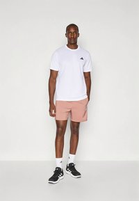 White short-sleeve t-shirt with black logo, paired with pink athletic shorts and black sneakers. Model stands on a neutral background.