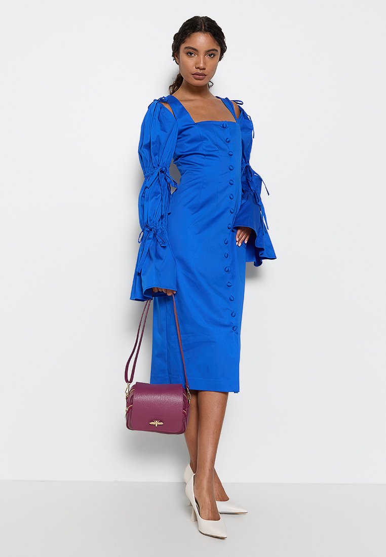Vestido midi azul royal con mangas largas abullonadas y detalles atados, con un cierre frontal de botones. Combinado con un bolso burdeos y tacones blancos.