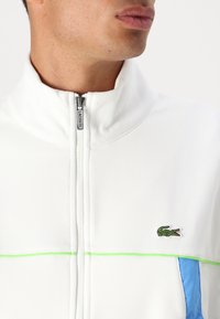 Balta zip-up striukė su žaliais ir mėlynais akcentais. Pasižymi tekstūruotu audiniu, aukštu apykakle ir Lacoste logotipu kairėje pusėje.