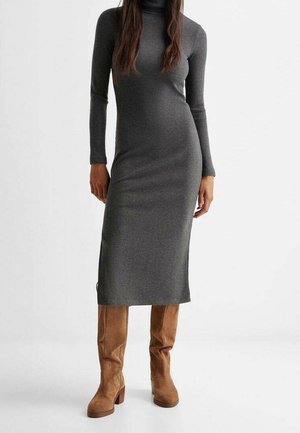 Femme portant une robe midi grise côtelée ajustée à manches longues avec un col roulé et des bottes en daim marron montant jusqu'aux genoux, debout sur un fond blanc.