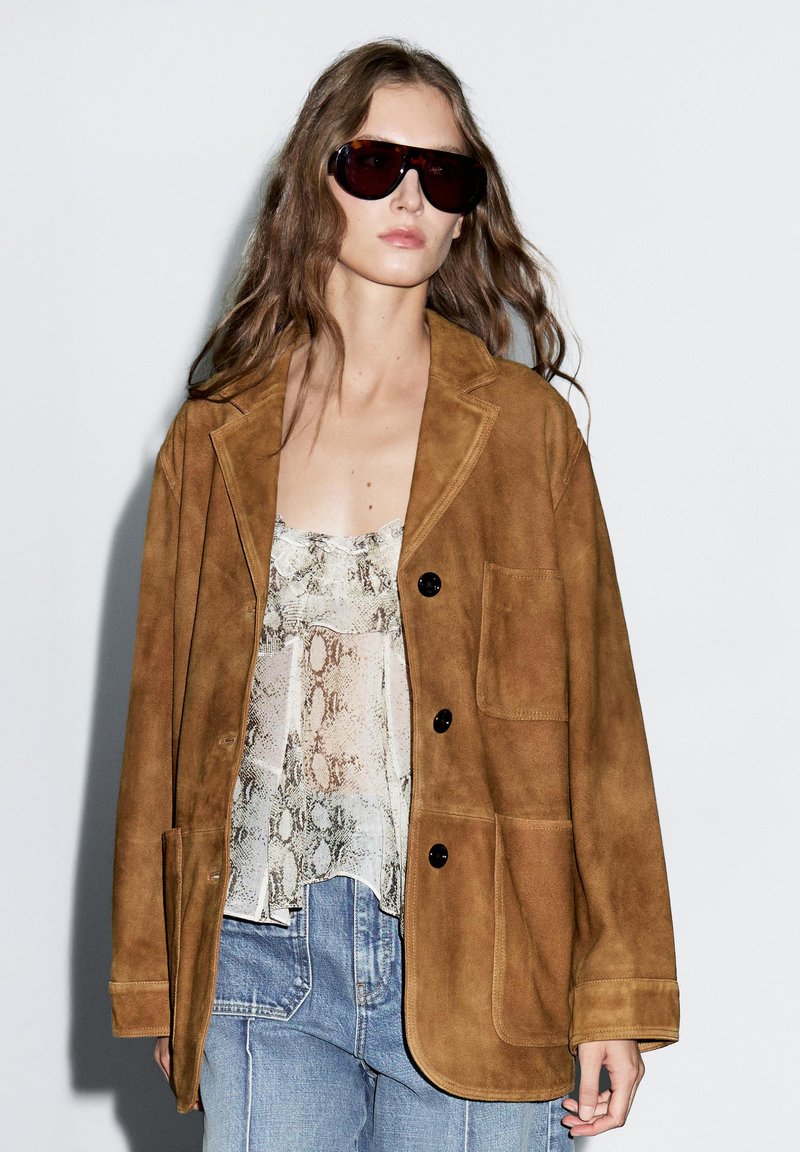 Giacca in suede marrone con colletto a svolta, tasca sul petto e bottoni neri, indossata sopra una maglietta con stampa a serpente e jeans azzurri chiari.