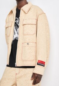 Beige jacka med ett texturerat, upprepande logomönster, kontrasterande röda sömmar, två framfickor och ett varumärkespatch på ärmen.
