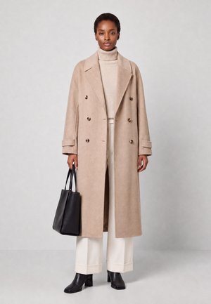 Vrouw die een beige dubbelgeknoopt lange jas draagt, een crèmekleurige coltrui, witte broek met wijde pijpen, zwarte laarzen en een zwarte handtas vasthoudt.