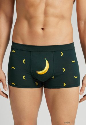 Pantalones cortos de boxeo verde oscuro hechos de algodón, con estampados de plátanos amarillos de diferentes tamaños, incluyendo un plátano grande en la parte delantera.