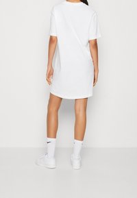 Bílé oversized tričko šaty, bavlněný materiál, kulatý výstřih, krátké rukávy, jednoduchý design. Nosené s bílými sneakers a ponožkami s černým logem.