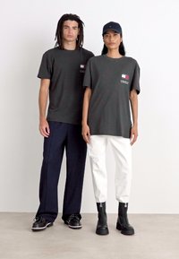 Dos modelos llevan camisetas oversized: una en gris oscuro con un logo y la otra en blanco, combinada con botas negras y pantalones azul marino.