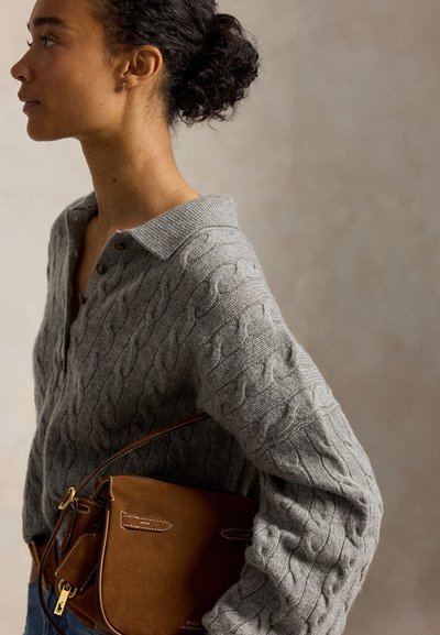 Γυναίκα με σγουρά μαλλιά και γκρι πλεκτό πουλόβερ κρατά καφέ δερμάτινη τσάντα μπροστά σε απλό φόντο.