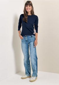 Navy long-sleeve shirt with contrasting side panels, paired with light blue straight-leg jeans and beige sneakers. Simple, functional design.