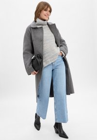 Vrouw in een grijze jas, gestreepte turtleneck trui, wijd uitlopende lichtblauwe jeans, zwarte hakken, met een kleine zwarte handtas.