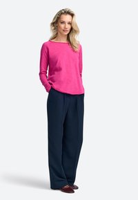 Rosa Langarmshirt mit weitem Ausschnitt, kombiniert mit dunkelblauen Weitbein-Hosen und bordeauxroten Slippers. Glatte Texturen und lässige Passform.