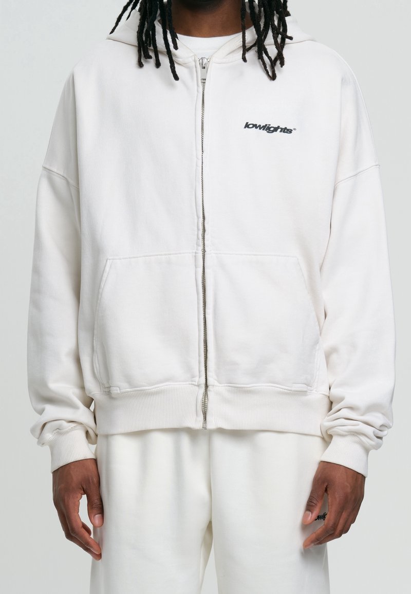 Homme portant un sweat à capuche blanc surdimensionné avec fermeture éclair et poches avant, arborant le logo "lowlights" sur la poitrine, assorti à un pantalon blanc.