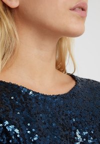 Blondi henkilö, jolla on tummansininen paljetti-paita pyöreällä kaula-aukolla, näkyy alhaalla oleva kasvojen osa, kaula ja olkapää.