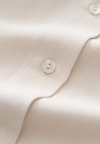 Light beige linen fabric with a subtle texture, featuring a single round button near the edge. Smooth finish and sewn seam details.