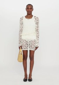 Rochie albă din croșetat cu mâneci lungi, având un model floral decupat. Împerecheată cu o geantă mică din fibră de iarbă bej și pantofi negri.