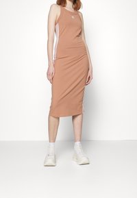 Brun figurnära klänning med ärmlös design, vita sidosträck och Adidas-logotyp. Bärs med ljusfärgade chunky sneakers.