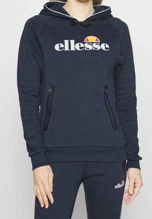Person wearing navy blue ellesse hoodie and matching sweatpants with front zip pockets and logo on chest and thigh.