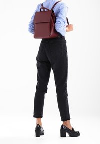 Femme portant un jean taille haute noir et des mocassins noirs à talons, avec un sac à dos en cuir rouge foncé, vêtue d'une chemise rayée bleue.