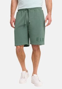 Grüne Baumwoll-Shorts mit einem elastischen Bund, Kordelzug und dem aufgedruckten "ELB" am unteren linken Bein. Kombiniert mit weißen Sneakers.