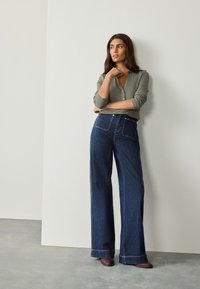 Top verde oliva con bottoni abbinato a jeans scuri a vita alta e gamba larga. Scarpe marroni con tacco basso completano l'outfit.