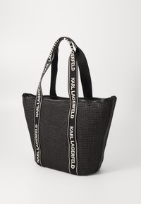 Black woven tote bag with branded straps, flat base, and zip top closure. Features textured surface and contrasting strap detailing.