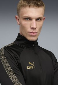 Svart atletisk kvart dragkedja topp med guldaccent, som har ett geometriskt mönster på ärmarna och logo-texten "KING" i guld.