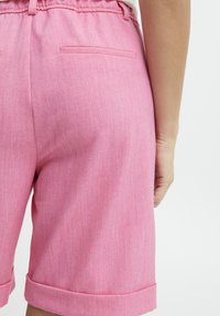 Vaaleanpunaiset shortsit, jotka on valmistettu pehmeästä ja teksturoidusta kankaasta. Ominaisuuksiin kuuluvat litteä vyötärö, käännetyt lahkeet ja takatasku.