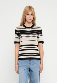 Jeune femme aux longs cheveux blonds portant un pull à manches courtes rayé beige et noir et un jean bleu clair sur fond blanc.