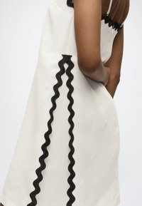 Γυναίκα με λευκό αμάνικο φόρεμα με μαύρες κυματιστές λεπτομέρειες στην πλάτη και στα πλάγια, όρθια με τα χέρια λυγισμένα πίσω.