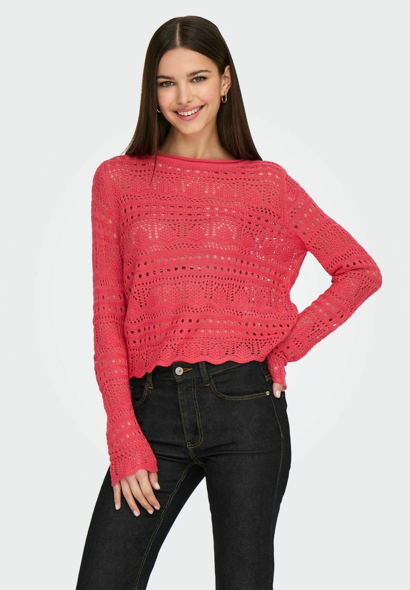 Pull en crochet de couleur corail vif, avec un motif texturé, des manches longues et un ourlet court, associé à un jean foncé.