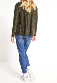 Oliivinvihreä neulepaita rentoa mitoitusta, yhdistettynä vaaleansinisiin farkkuihin. Valkoiset tennarit mustilla yksityiskohdilla täydentävät asukokonaisuutta.