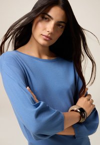 Pull en tricot bleu avec un large col et des manches trois-quarts, porté par une personne aux cheveux sombres et longs. Deux bracelets empilés au poignet.