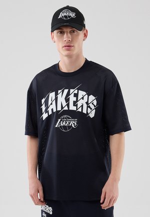Νεαρός άνδρας που φοράει μαύρο καπέλο Los Angeles Lakers, μαύρο διχτυωτό μπλουζάκι Lakers και ταιριαστά σορτς, στέκεται μπροστά σε απλό γκρι φόντο.