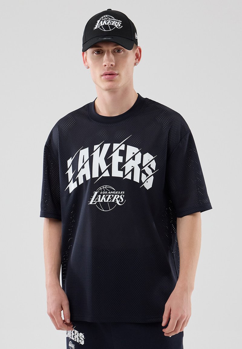 Νεαρός άνδρας που φοράει μαύρο καπέλο Los Angeles Lakers, μαύρο διχτυωτό μπλουζάκι Lakers και ταιριαστά σορτς, στέκεται μπροστά σε απλό γκρι φόντο.