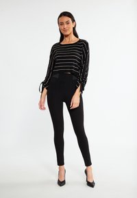 Zwart cropped top met gouden horizontale strepen en strikdetails bij de mouwen, gecombineerd met hoge zwarte leggings en zwarte hoge hakken.
