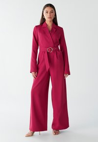 Fuksijasti kombinezon z dolgimi rokavi, ovratnikom v slogu blazerja in pasom s pasom. Dizajn z širokimi hlačnicami in gladko teksturo. Zlati dodatki.