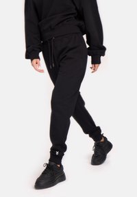 Melnas sporta bikses, kas izgatavotas no mīksta materiāla, ar elastīgu jostu un auklu, ribotām piedurknēm un mazu logotipu uz kreisās potītes.