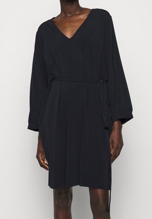 Personne portant une robe portefeuille noire à manches longues avec un col en V et une ceinture nouée à la taille sur un fond clair uni.