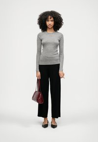 Γκρι ριγέ μπλούζα με μακριά μανίκια, μαύρο παντελόνι σε φαρδιά γραμμή, μαύρες μπαλαρίνες και μπορντό τσάντα, με σγουρά μαλλιά, να στέκεται μπροστά σε ουδέτερο φόντο.