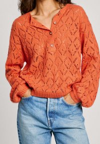 Texturerad, stickad orange tröja med ett diamantmönster, en knäppen henley-halsringning och ribbade ärmslut, i kombination med ljusblå jeans.