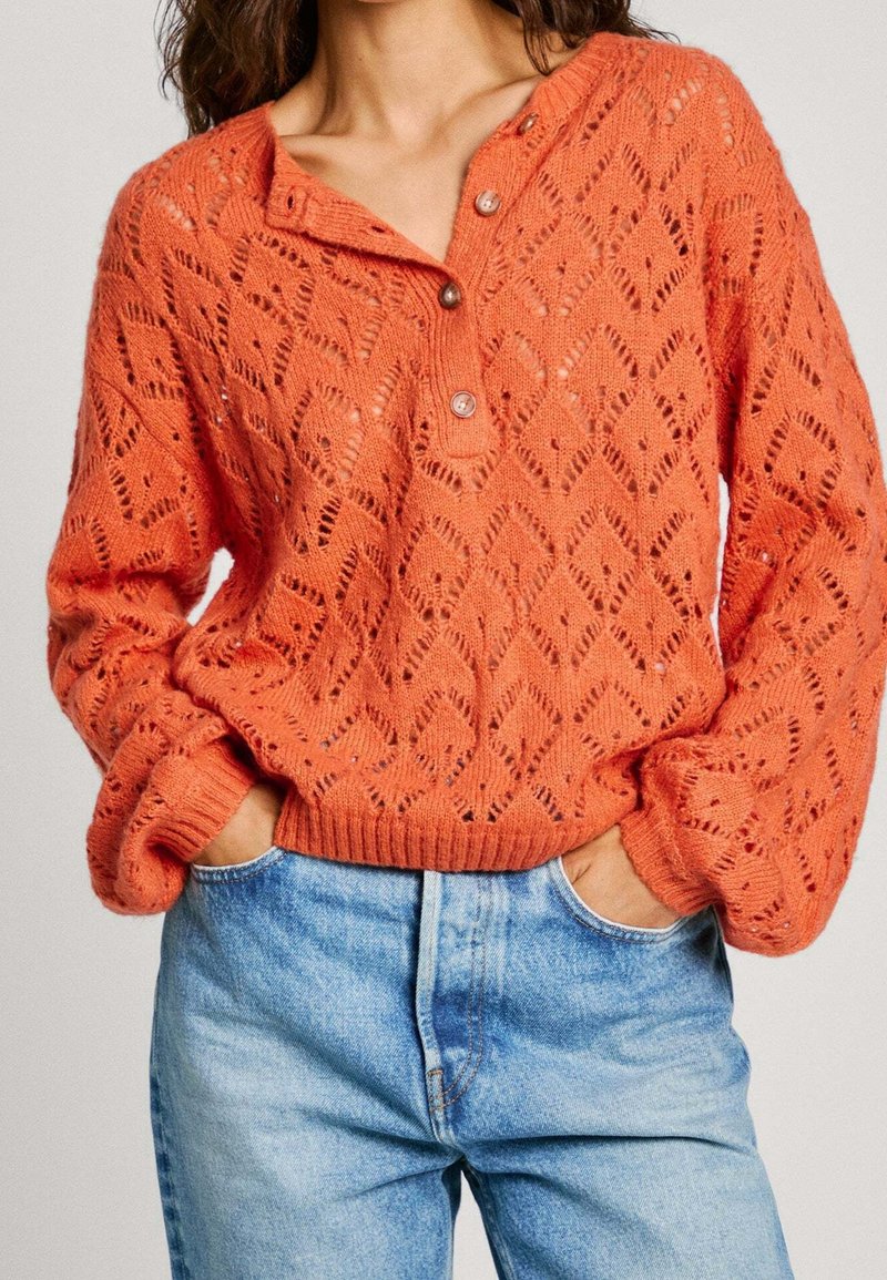Texturerad, stickad orange tröja med ett diamantmönster, en knäppen henley-halsringning och ribbade ärmslut, i kombination med ljusblå jeans.