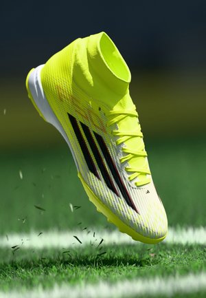 Geltoni ir balti futbolo bateliai su aukštu čiurnos dizainu, juodom juostelėm ir tekstūruotu paviršiumi, pakelti virš žalios žolės.