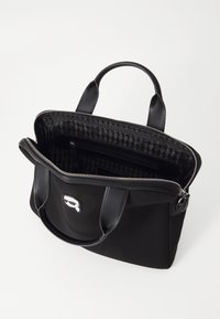 Czarna torba typu tote z zamkiem błyskawicznym i podwójnymi uchwytami. Wnętrze wyłożone wzorzystą podszewką z kieszenią na zamek. Na zewnętrznej stronie widoczny jest akcent z logo.