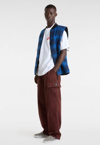 Blauwe geruite vest over een wit T-shirt met rood logo, gecombineerd met losse bordeauxkleurige cargobroeken en zwart-wit geruite instap schoenen.