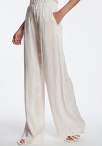 Lichtbeige wijde broek van textuurstof. Beschikt over een elastische tailleband en zijzakken. Soepele, vloeiende uitstraling met een subtiele glans.