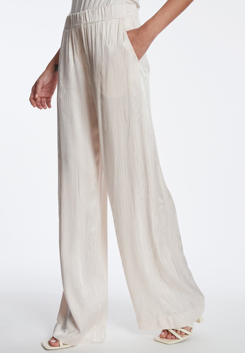 Lichtbeige wijde broek van textuurstof. Beschikt over een elastische tailleband en zijzakken. Soepele, vloeiende uitstraling met een subtiele glans.