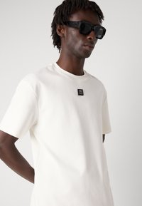 Cremfarbener Kurzarm-T-Shirt mit rundem Ausschnitt und einem schwarzen Logo-Patch auf der Brust. Der Stoff wirkt glatt mit einem leichten Glanz.