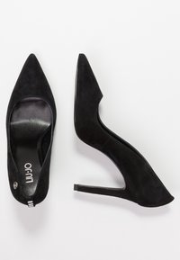 Zapatos de tacón alto de ante negro con puntera puntiaguda y lados recortados. Presentan un tacón delgado y el logo en el interior. Diseño sencillo sin acentos adicionales.